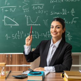 impressed young female teacher sits at table with school tools raising pen in classroom